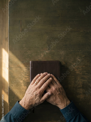 Man's hands resting on a closed leather Bible over a rustic wooden table