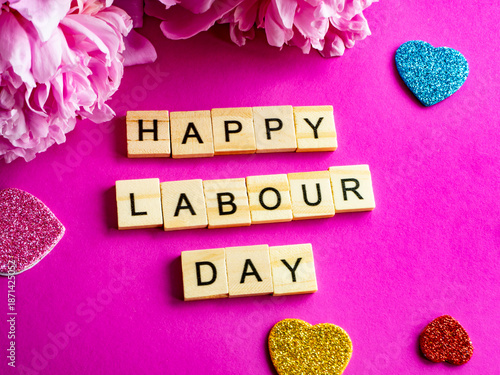 wooden blocks spelling happy labour day on pink background, pink peony and colourful hearts