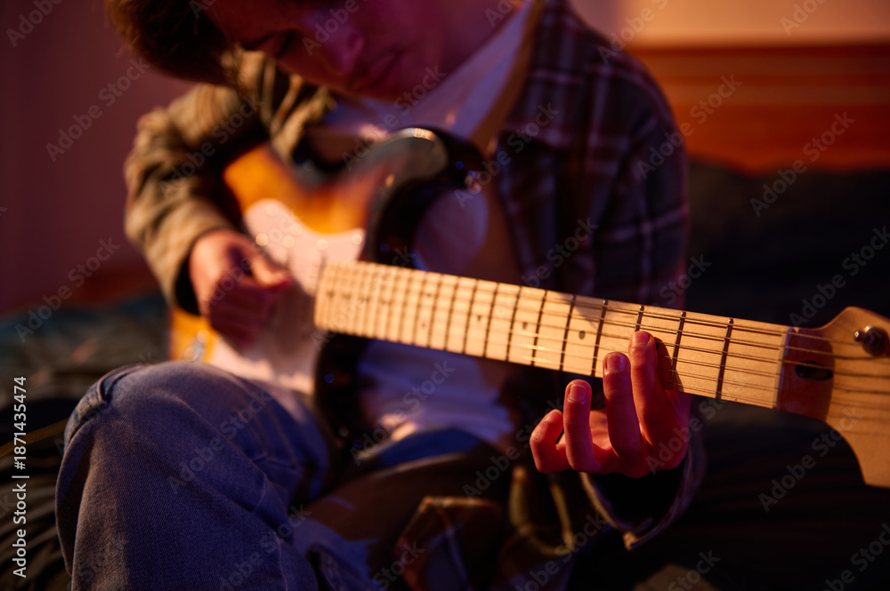 Fototapeta premium A guitarist sits indoors in warm amber light, fingers on the fretboard, enjoying a focused, intimate moment of practice and music.