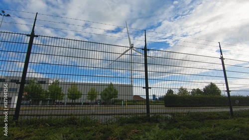 Wind turbine behind metal security fence in an industrial area, renewable energy infrastructure with modern technology, cloudy sky with blue patches, daytime outdoor scene, no people