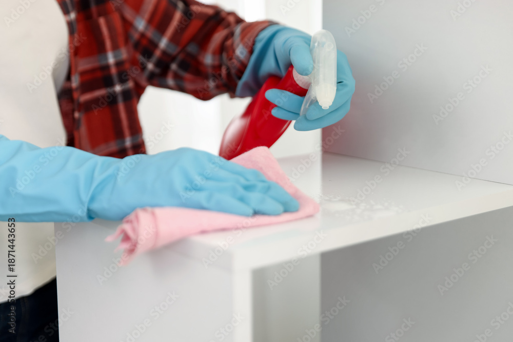 Fototapeta premium Woman spraying cleaning product while wiping dust from shelving unit indoors, closeup