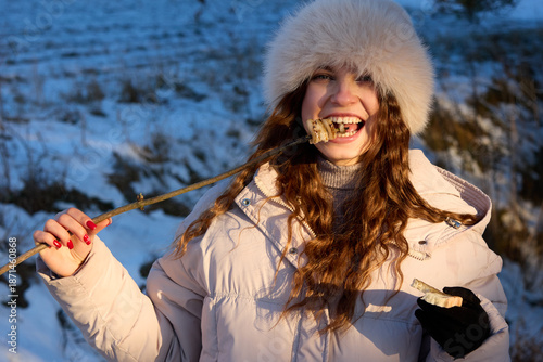 A happy young woman dressed in winter attire savoring a piece of toasted marshmallow on a stick outdoors against a snowy landscape during the winter season.