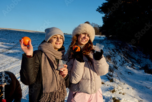 Two happy young women enjoying a sunny winter day outdoors, holding oranges and cups, dressed warmly in stylish hats and scarves, sharing a joyful moment together in a snowy landscape.