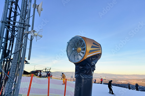 Snowmaking machine on ski slope with clear blue sky and people skiing