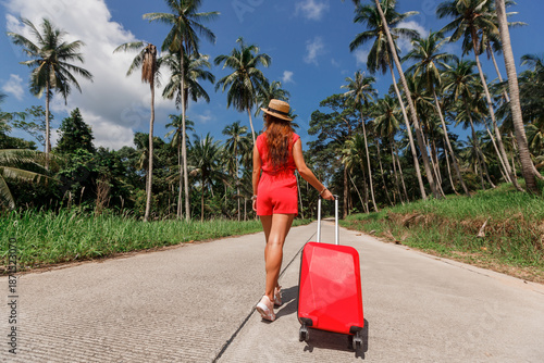 Attractive girl in summer clothes on a tropical island by the sea with a red suitcase, travel, searching for a hotel, searching for transport, waiting for a transfer, summer mood