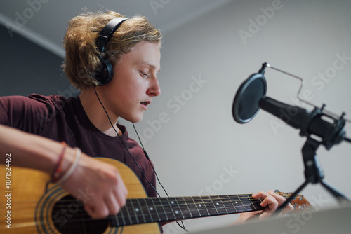 Bild auf Leinwand Home sound studio portrait of young teenager playing guitar in headphones, recording voice and music using microphone and laptop
