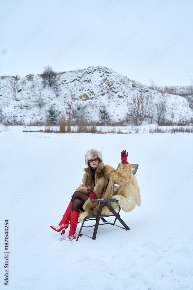 Fototapeta premium Stylish woman in fur coat, hat, and red boots sits on a chair in a snowy field with arm raised, creating a visually interesting contrast against the winter backdrop.