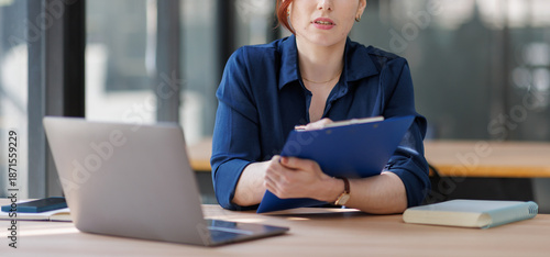 Middle aged serious woman working with laptop sitting at table make notes in copybook at office
