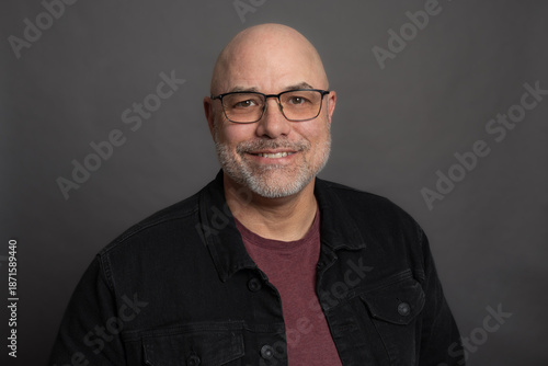 Mature, bald man with beard and glasses wearing black jacket, isolated on grey background