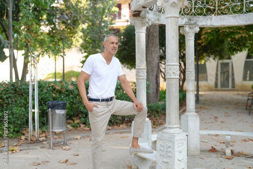 A man in a white shirt and tan pants is standing in front of a stone archway. He is stretching or doing some sort of exercise. The scene is set in a park or courtyard, with trees
