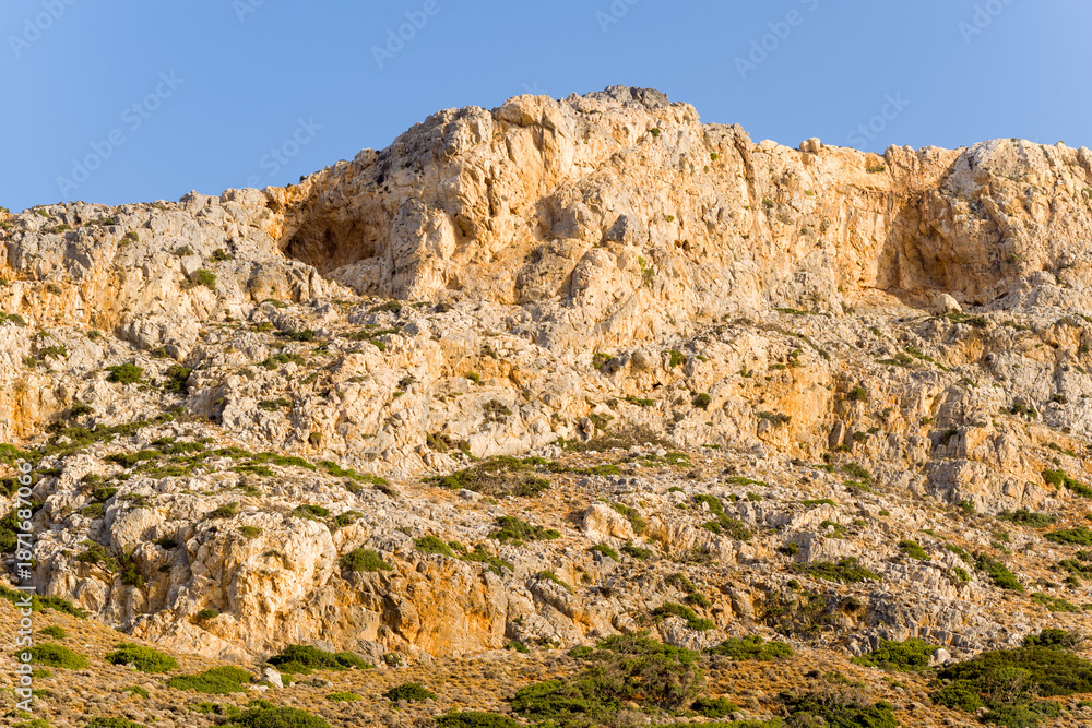 Fototapeta premium Warm sunlight highlights the rugged textures and earthy tones of a rocky cliff face in the mountains near Balos, Crete. Sparse vegetation is scattered across the jagged limestone surface under a clear