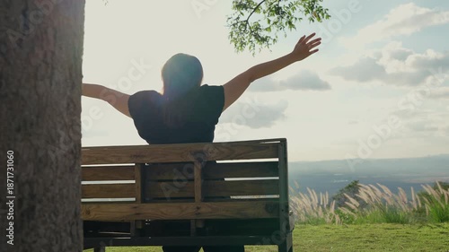 Rear view of a woman sitting alone on a wooden bench under a large tree, enjoying the mountain view, breathing in fresh air, and immersed in a peaceful natural atmosphere