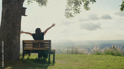 Rear view of a woman sitting alone on a wooden bench under a large tree, enjoying the mountain view, breathing in fresh air, and immersed in a peaceful natural atmosphere