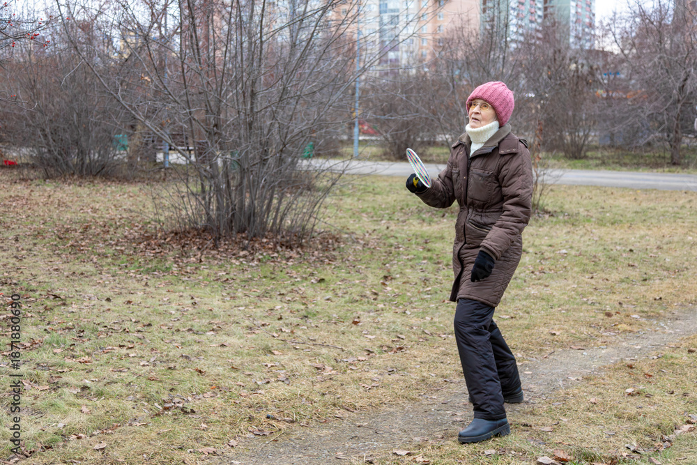 Naklejka premium Elderly woman in winter clothes playing badminton in an autumn park. Active senior lifestyle, outdoor recreation for healthy living.