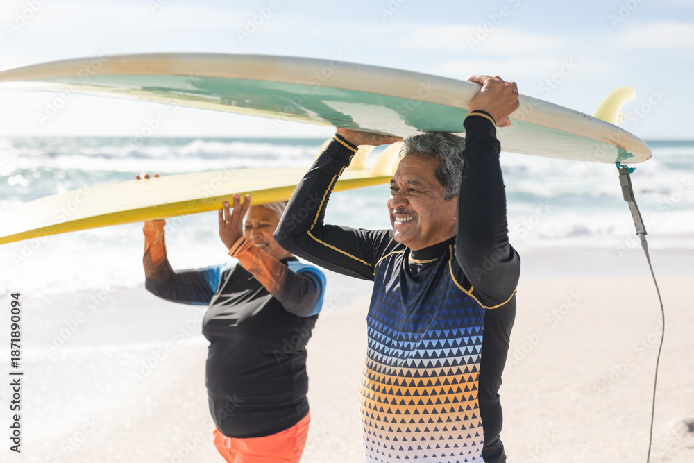 Fototapeta premium Happy multiracial senior couple carrying surfboards over heads at beach during sunny day