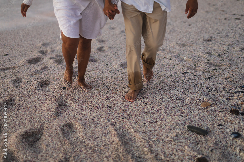Fototapeta premium Low section of senior multiracial couple holding hands walking barefoot on sand at beach