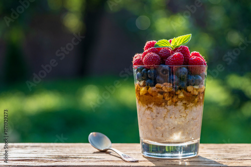 Summer breakfast layered dessert in a glass of oatmeal, blueberries, raspberry, walnut, olive oil and honey on nature background, closeup. Healthy diet with fresh berries on wooden table