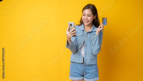 Smiling Asian Woman Holding Credit Card and Smartphone in Studio with Bright Yellow Background, Young Female Making Online Payment, Digital Banking, E-Commerce, and Cashless Lifestyle Concept