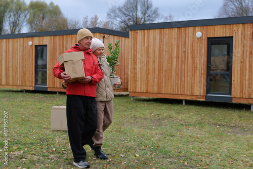 Obraz na plátně An older couple carries boxes and a plant while walking in front of wooden cabin