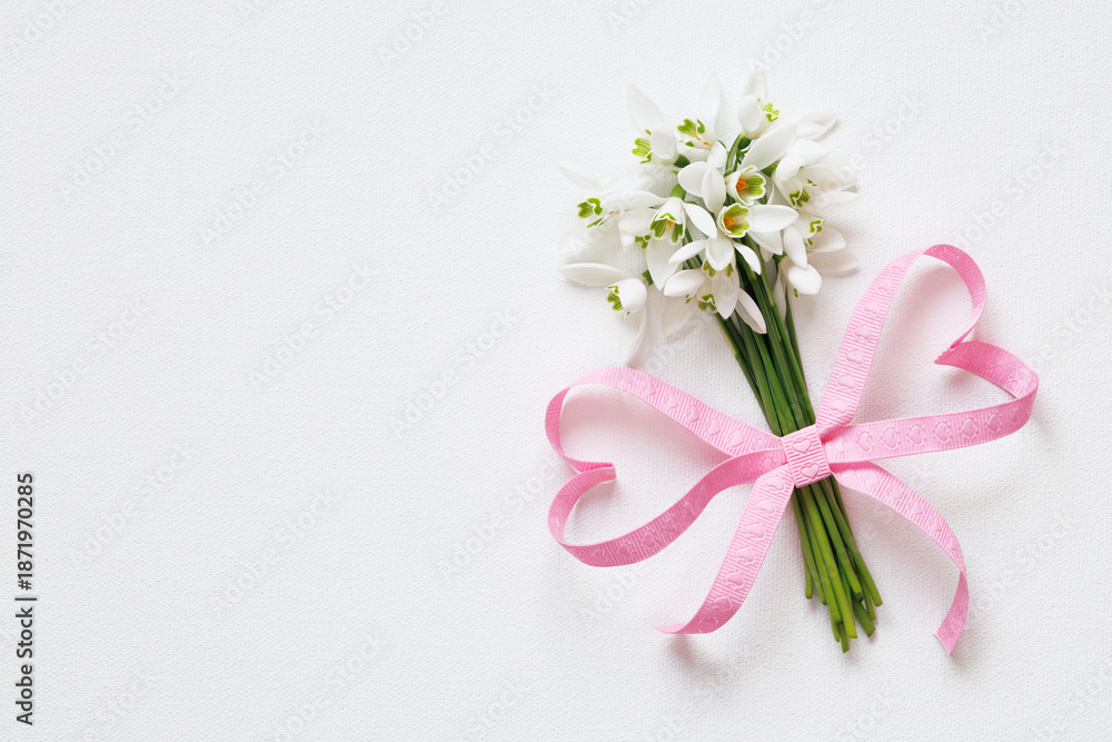 Fototapeta premium Bouquet of snowdrop flowers with a bow of pink ribbon in the shape of hearts on a white background. Beautiful postcard, copy space