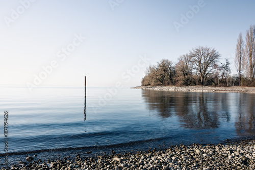 Ruhiges Seeufer mit Kieselstrand und Markierungspfahl im Wasser