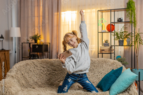 Young teenager girl takes moms phone at home, giggling while playing passing a tough online game level with excitement. Child kid on sofa shouting i did it and waving phone high in joy, celebrate win