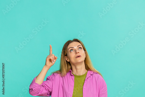 Middle-aged woman points finger upward with serious face, clearly signaling importance, asking to focus attention on something above. Girl isolated on blue background shows emphasis gesture and intent
