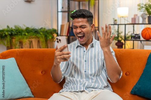 Indian man at home scrolls smartphone feed sees sale news and says wow smiles enjoy discount deals. Arabian guy on sofa reads offer plans order adds item to cart and feels good reaction to price drop.