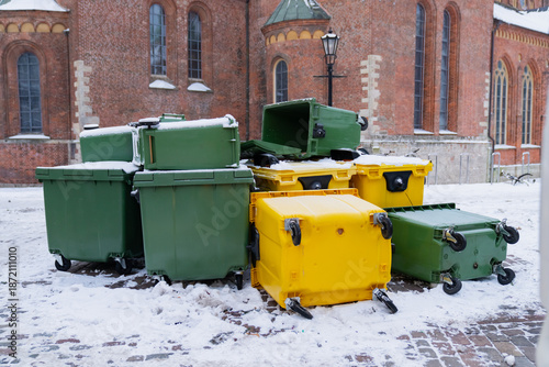 Fallen garbage containers with wheeled green and yellow bins lying on snowy street surface showing urban waste management disruption and cold season conditions
