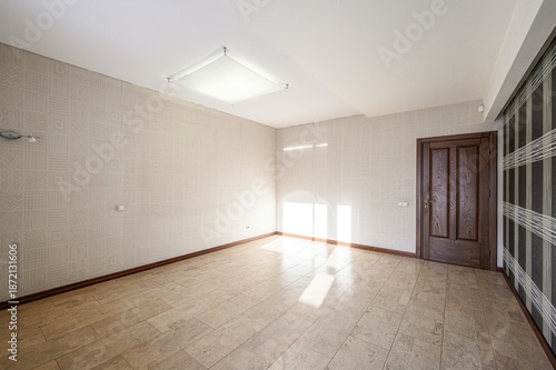 Spacious unfurnished room with beige patterned wallpaper, tiled floor, wooden door, recessed ceiling light, natural light from window, dark paneled section on one wall. Clean, modern look