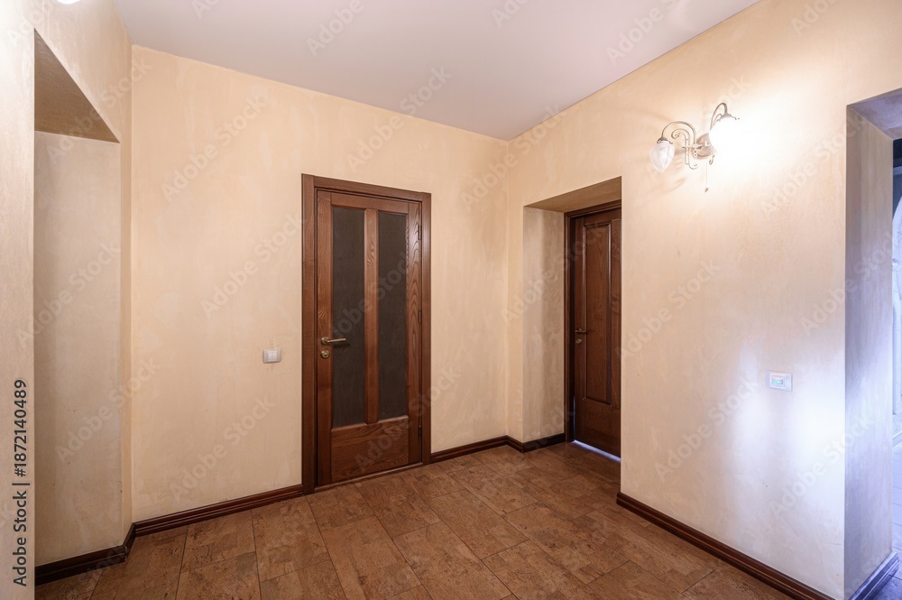 Obraz premium A minimalist hallway with beige textured walls, brown tiled floor, wooden doors (one with frosted glass panels), and wall-mounted sconces providing soft lighting. Clean, uncluttered design