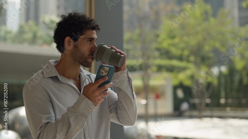 Portrait of young Hispanic man holds smartphone and drinks coffee in urban area slow motion. Smiling Latin male reads message as walks past panoramic window in apartment complex