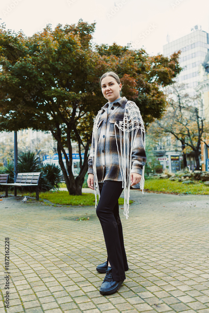 Fototapeta premium Young woman stands in park wearing a checkered coat with fringe, surrounded by trees and benches during a cloudy day