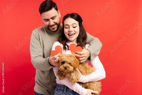 Smiling happy woman and man holding pretty dog and red heart hugging isolated on red background