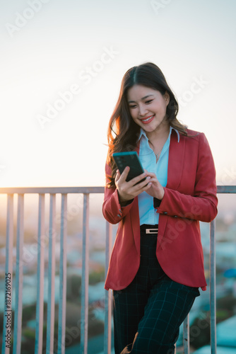Young Asian businesswoman in a red blazer smiling and texting on her mobile phone. Leaning against a railing on a city rooftop with a warm sunset glow creating a professional and connected atmosphere