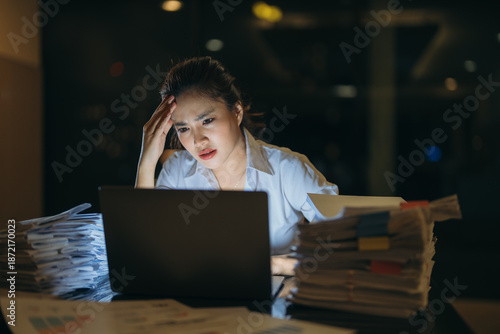 Young woman working overtime feeling stressed and exhausted from a heavy workload and tight deadlines, experiencing a headache while looking at her laptop in a dark office at night