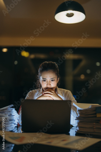 Young businesswoman feeling overwhelmed and exhausted, focusing intently on her laptop screen late at night, surrounded by stacks of paperwork and facing a demanding deadline