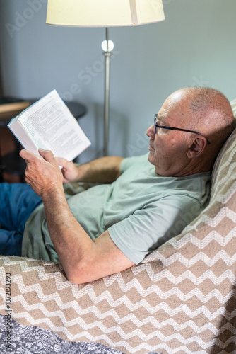 An older man is reading a book while comfortably seated on a cozy chair at home. The warm light from a nearby lamp adds to the peaceful atmosphere of the afternoon.