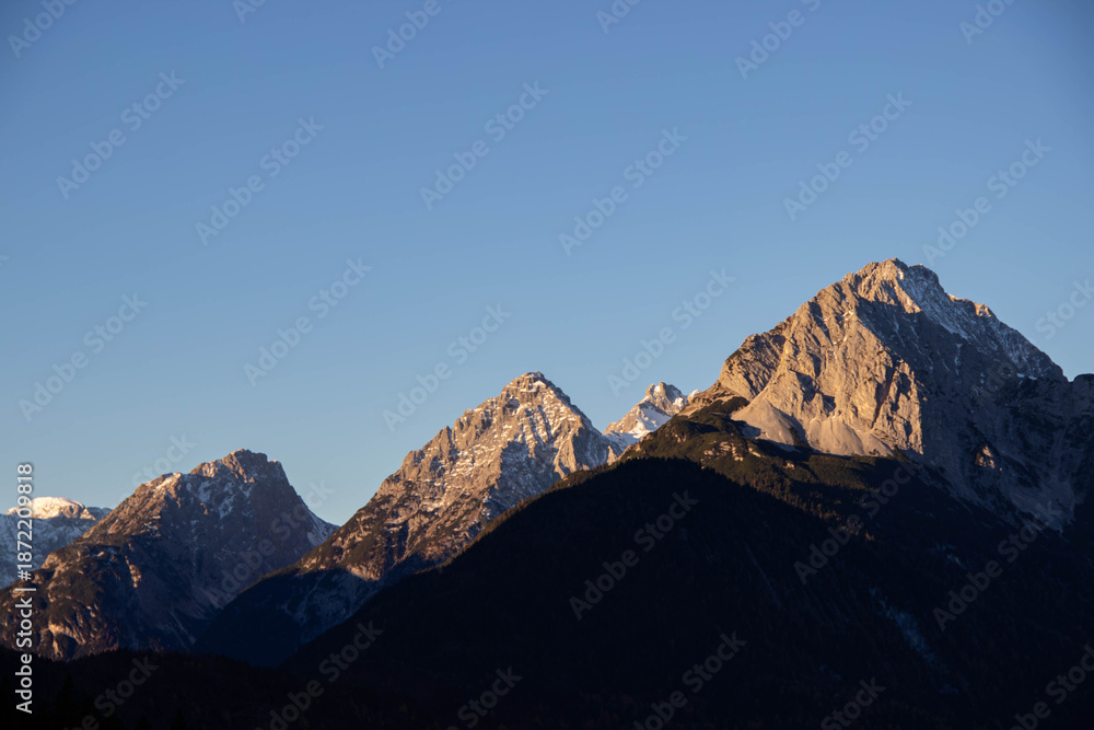 Fototapeta premium Majestic mountain peaks illuminated by golden sunlight, showcasing rugged terrain and snow-capped summits against a clear blue sky, embodying the beauty of nature's landscape