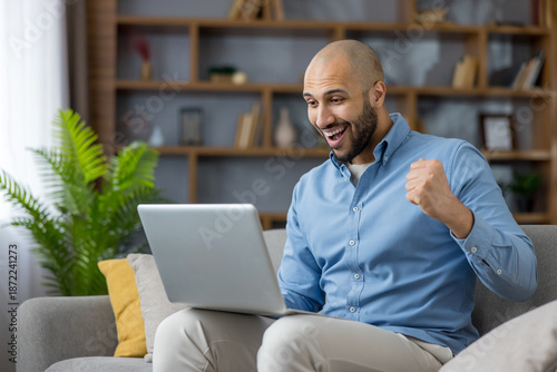 Foto Happy young man sitting on a sofa, looking at his laptop screen, raising his fis