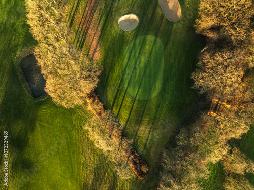 Aerial drone view of golf course green at golden hour with long tree shadows and pond
