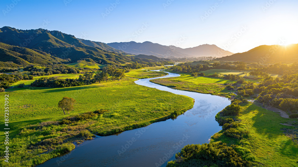 Fototapeta premium Serene landscape featuring winding river surrounded by lush green fields and mountains clear blue sky. warm sunlight creates peaceful atmosphere