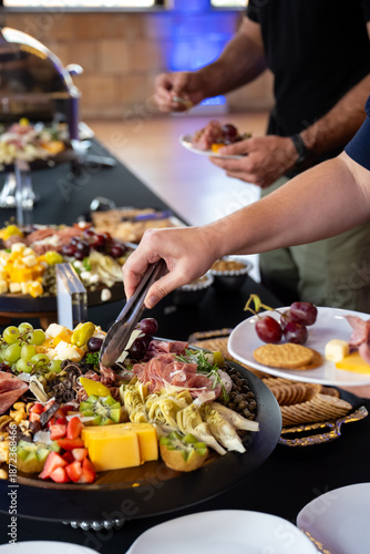 People selecting food from a catered appetizer table featuring cheese, fruit, cured meats, and crackers during a social gathering.
