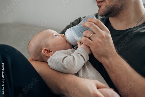 Canvas-taulu A father feeds his newborn son from a bottle of milk while sitting on the bed