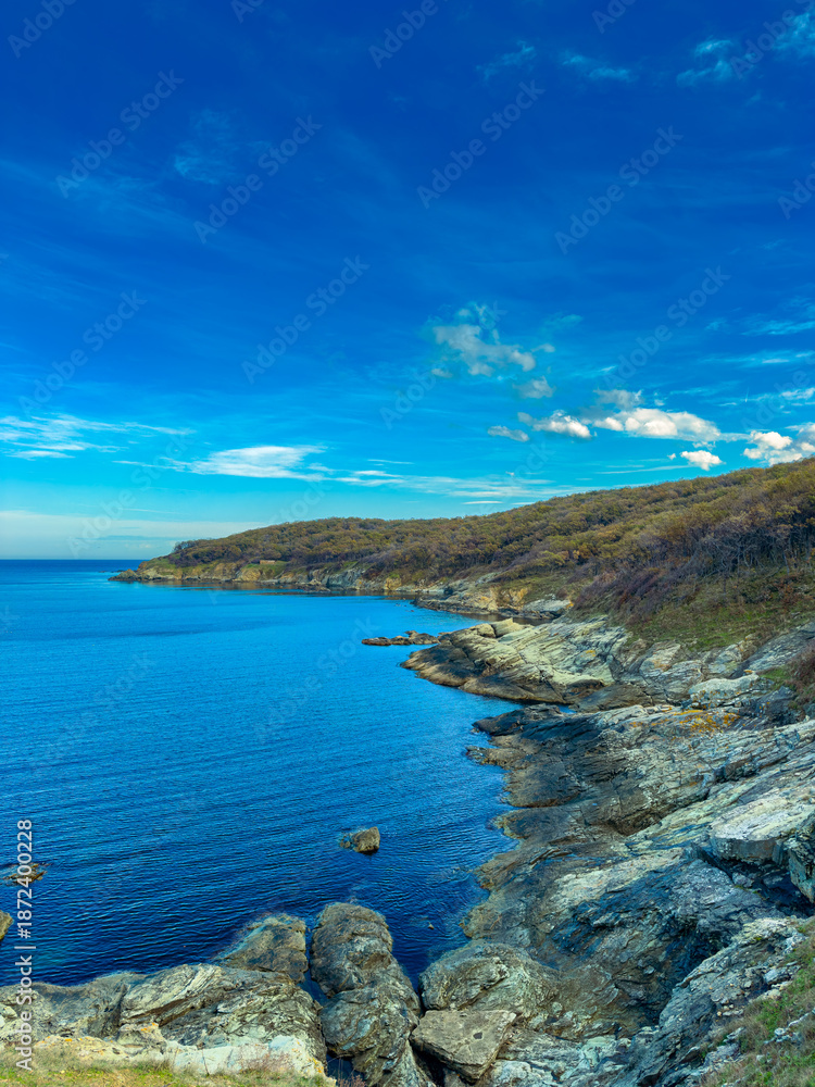 Fototapeta premium Clear sky over rocky coastline with calm water at midday near a green hillside