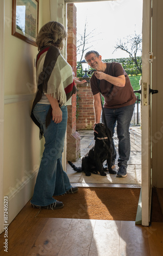 Dog walker returning a black cocker spaniel after his walk, exercise. A woman pet owner takes the lead from the man at the doorstep, view from inside of the hallway of the house.