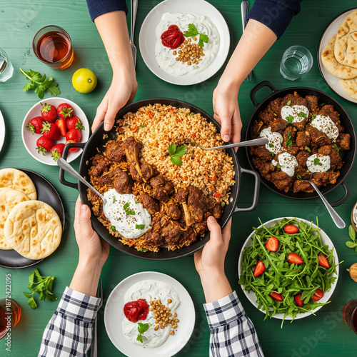 Family having Turkish dinner. Flat-lay of people passing rice pilav over green table with lamb in yogurt sauce, fresh arugula and strawberry salad and flatbread, top view. Ramazan iftar supper