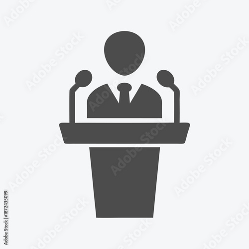 A speaker wearing a tie is standing at a podium with two microphones