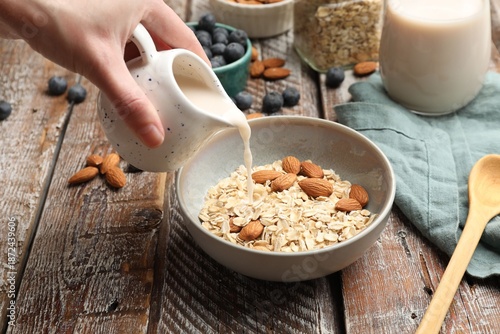 Photos Woman pouring almond milk into bowl with oat flakes and nuts at wooden table, cl