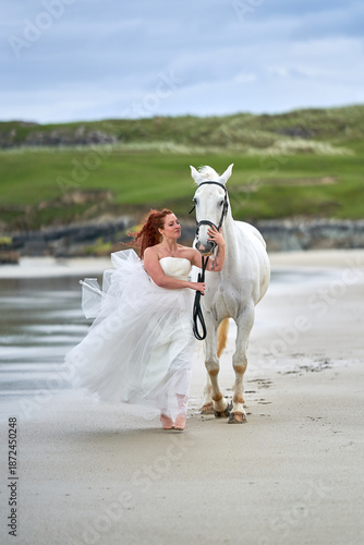 A white horse and a woman in a wedding dress stand on a beach, with the sky and sea.